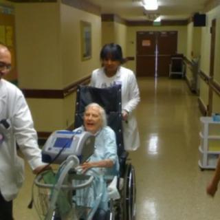 Healthcare workers assist an elderly woman in a wheelchair down a hospital corridor.