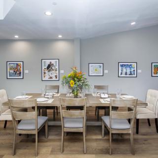 A contemporary dining room featuring a large wooden table, surrounded by chairs and adorned with vibrant flower arrangements. The walls are decorated with framed artworks.