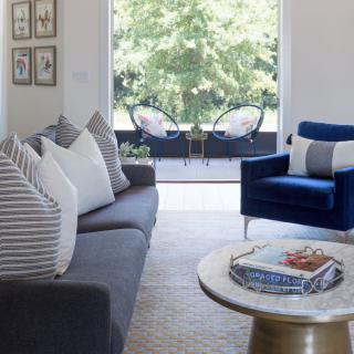 A modern living room with a stylish blue armchair, striped pillows on a dark sofa, and a round coffee table overlooking an outdoor patio.