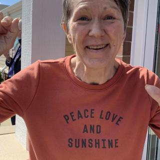 A person smiling while wearing a shirt with the message 'Peace, Love, and Sunshine' outdoors.