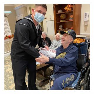 A masked military personnel interacts with an elderly veteran in a wheelchair indoors.
