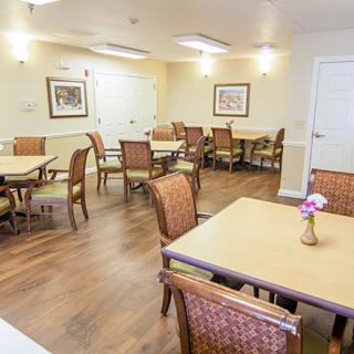 A well-lit dining room with neatly arranged tables and chairs, featuring wood flooring and decorative artwork on the walls.