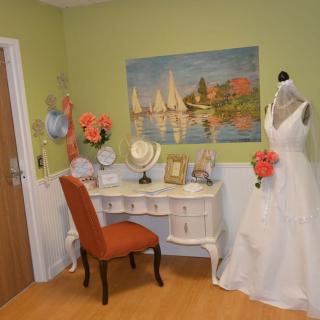 A charming bridal room featuring a wedding dress on a mannequin, a vanity table with an upholstered chair, floral decorations, and a sailboat painting on the wall.
