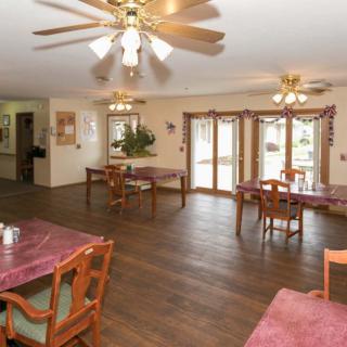 A spacious dining room with wooden floors, multiple tables and chairs, and ceiling fans, well-lit by natural light from the windows.