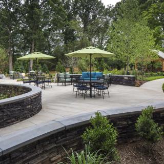 A landscaped outdoor patio area with tables, chairs, and green umbrellas surrounded by a stone wall and greenery.