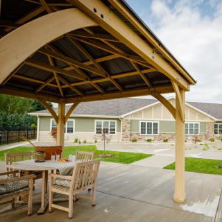 A cozy outdoor gazebo with wooden furniture on a patio, set against the backdrop of a modern single-story building.