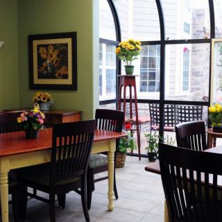 A cozy dining area with wooden tables and chairs, featuring floral decorations and large windows that let in natural light.