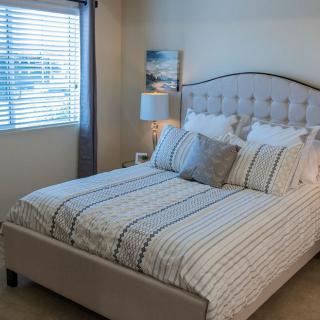 A cozy bedroom with a neatly made bed featuring striped bedding, a bedside lamp, and a window with blinds and curtains.