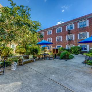 A peaceful courtyard with benches, trees, and blue umbrellas in front of a brick building under a clear sky.