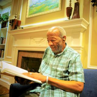 An elderly man enjoying reading a book by the fireplace.