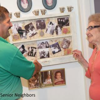 A senior woman and a man share a moment while looking at family photos on the wall.