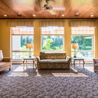 A cozy living room with ample natural light streaming through large windows, featuring comfortable seating and a detailed carpet.