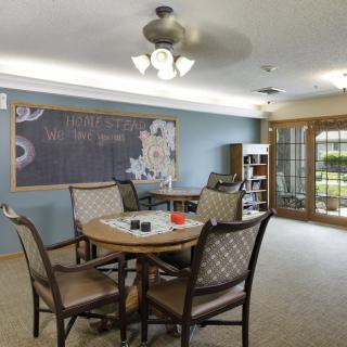 A cozy dining area with a central table and chairs, featuring a large chalkboard on the wall and natural light from the windows.
