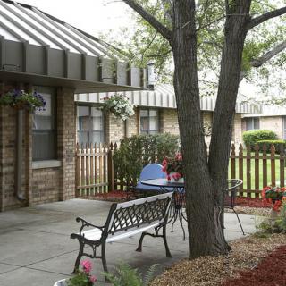 A peaceful courtyard features a bench and table set near a tree with surrounding fences.