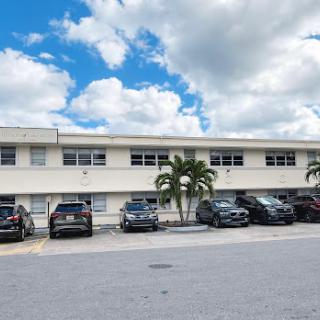 A view of a multi-story building with parked cars in front and palm trees under a cloudy sky.
