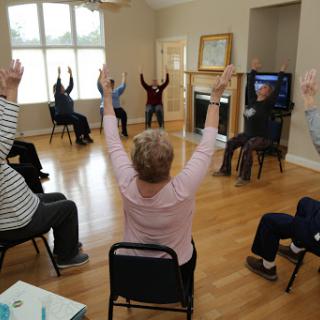 A group of seniors participating in a seated exercise class, raising their hands in unison.