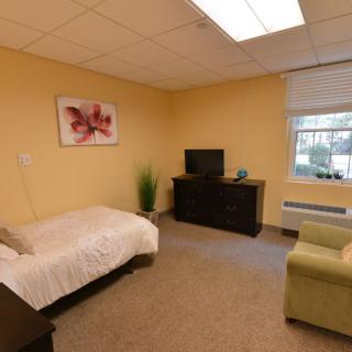 A cozy bedroom with warm lighting, featuring a bed, TV, chair, and window.