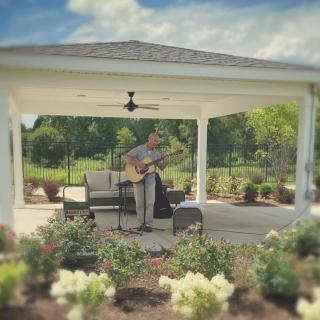 A musician playing guitar in a gazebo surrounded by a garden.