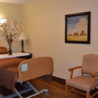 A cozy hospital room with a bed, chair, and decorative items including a painting and flowers.