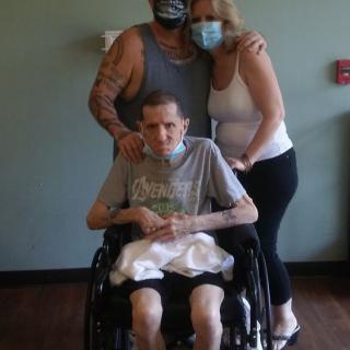 A group of three people, with one person in a wheelchair and the others standing behind them, all wearing masks in a healthcare setting.