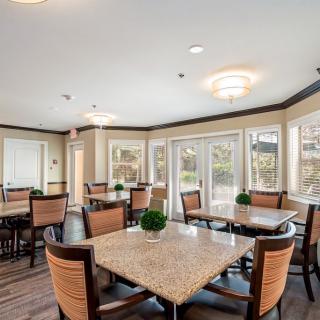 A well-lit dining area with tables and chairs, featuring large windows and indoor plants.