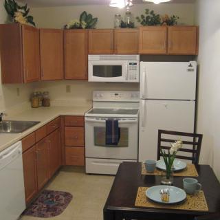 A modern kitchen with wooden cabinets, featuring a refrigerator, microwave, stove, and a small dining area.