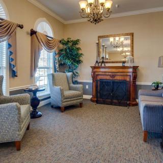 A classic, elegantly decorated living room featuring comfortable armchairs, a wooden fireplace with a decorative mirror above, and ambient lighting from a chandelier.