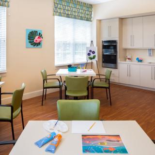 A bright art room with tables and chairs, featuring art supplies and a kitchen area.