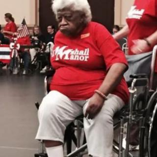 An elderly person in a red shirt sits in a wheelchair during an indoor event.
