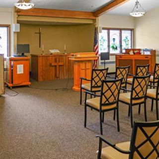 An empty chapel interior with rows of chairs facing a pulpit and cross.