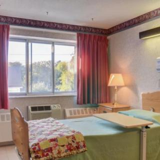 A hospital room with two beds, a large window with red curtains, and an adjacent table under warm lighting.
