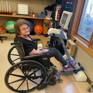 An elderly woman engaging in rehabilitation exercises using a specialized stationary bike while seated in her wheelchair.