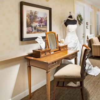 A vintage-style vanity setup featuring a mirror and chair, with a wedding dress on a mannequin in the background.
