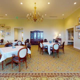A spacious dining room with elegant classic furniture and a central chandelier, featuring tables set with white tablecloths.