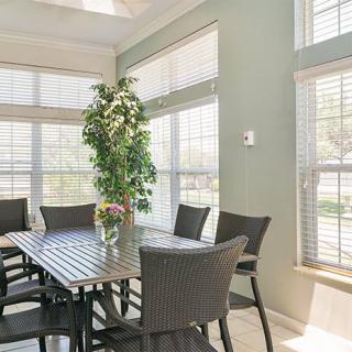 A bright dining room with a large table, surrounded by chairs and illuminated by natural light streaming through multiple windows with blinds.