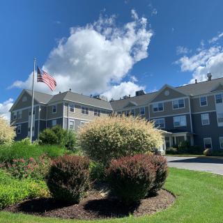 A scenic view of a building with an American flag, surrounded by landscaped bushes under a blue sky with clouds.