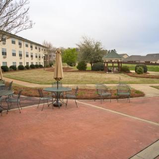 A courtyard with outdoor seating, surrounded by a pathway and trees, adjacent to a residential building.