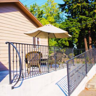 A sunny patio area with outdoor seating, an umbrella, and a railing, surrounded by trees.