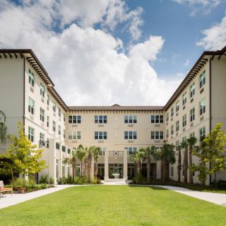 A four-story building with a central courtyard, framed by landscaped greenery and a partly cloudy sky.