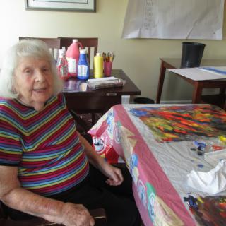 An elderly woman sitting by a table with art supplies, engaged in painting indoors.