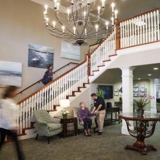 A spacious interior of a nursing home with a grand staircase, chandelier, and seating area where an assistant is helping a resident.