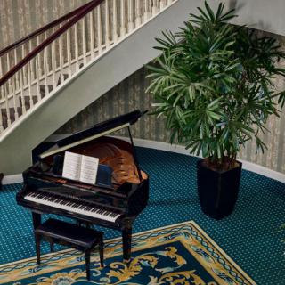 A grand piano with sheet music sits under a staircase next to a large potted plant, on a richly colored carpet.
