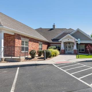 An exterior view of a building with a parking lot, featuring a brick facade and peaked roof.