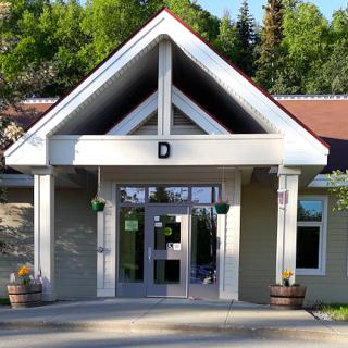 A building entrance with a gabled roof and glass doors, surrounded by greenery.