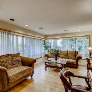 A spacious living room featuring comfortable sofas, wooden flooring, and large windows with vertical blinds.