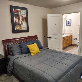 A cozy bedroom with a neatly made bed, featuring gray bedding and a yellow accent pillow. An open door leads to the bathroom.