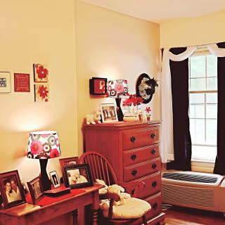 A cozy bedroom with warm decor, featuring a dresser and desk adorned with photos and a floral lampshade.
