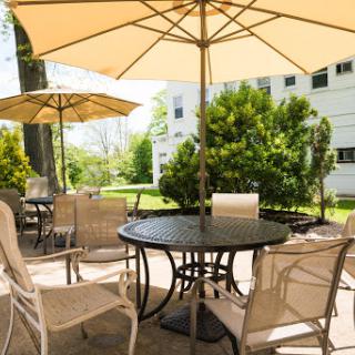 A serene outdoor patio setup with tables and chairs under large umbrellas, surrounded by lush greenery on a sunny day.