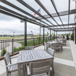 An outdoor patio area with tables and chairs under a pergola, overlooking a landscaped area with an American flag in the background.