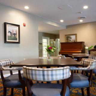 A cozy dining room featuring a wooden table surrounded by chairs, with a piano and framed artwork in the background.